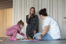 Stephanie met haar dochter spelend op de grond. Sandra filmt op de achtergrond.