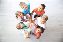 Zeven kinderen in een rondje met hun handen op elkaar kijkend naar boven naar de camera.